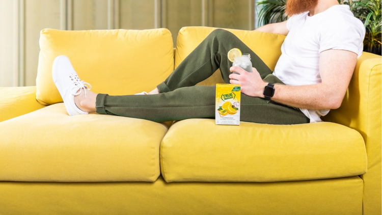 A man sitting on a big yellow couch drinking a class of water with True Lemon. 