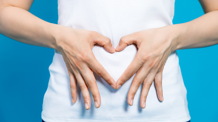 A person wearing a white shirt is standing in front of a blue background. Only their torso is shown, they are holding their fingers together in front of their stomach in the shape of a heart. 