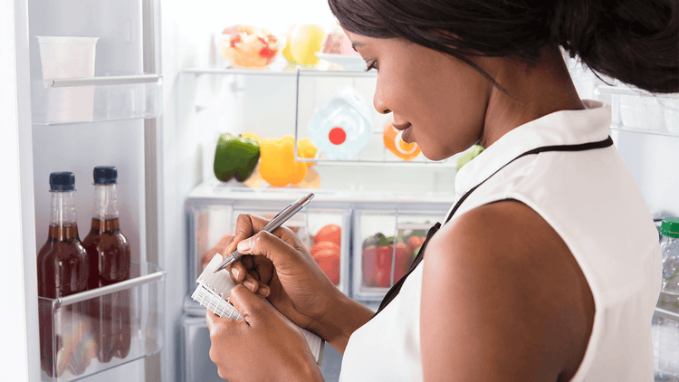 Woman stands in front of open refrigerator writing down a grocery list