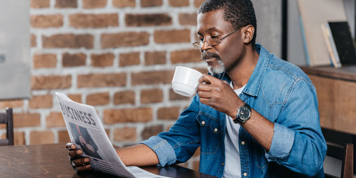 Man sits at a table drinking out of a white mug and reading a newspaper