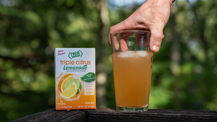 A glass of Triple Citrus Lemoande next to a box of Triple Citrus Lemonade are on a porch railing. There is a hand holding onto the glass of Triple Citrus.