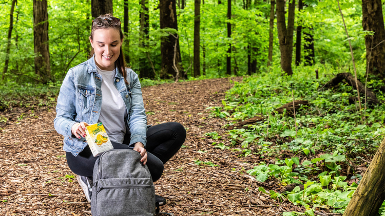 Woman in woods, placing box of True Lemon flavor packets in backpack.