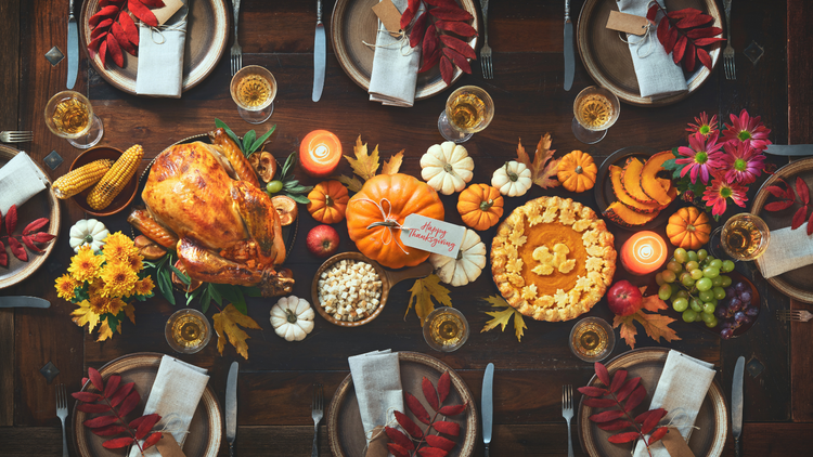A birds-eye view of a Thanksgiving spread. On the table there are eight plates, small pumpkins, orange and white candles, a golden turkey, a pumpkin pie, and some other small plates of food.  