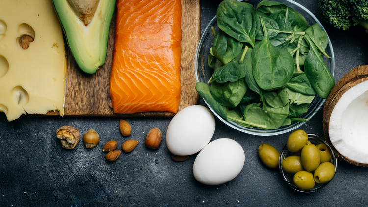 A birds-eye view of a spread of food. It includes uncooked salmon, an avocado sliced in half, two eggs, some nuts, slices of cheese, a bowl of spinach, half of a coconut, and olives.  