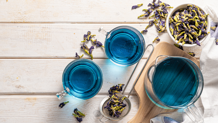 In the right side of the image are cups of steeped butterfly pea flower tea. The teal is a bright teal. Dried flowers are arranged around the cups.