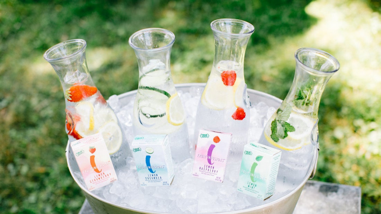 A tub of ice with glass pitchers filled with Fruit Infused water. Boxes of True Lemon Fruit Infusions sit in the ice in front of the pitchers. 