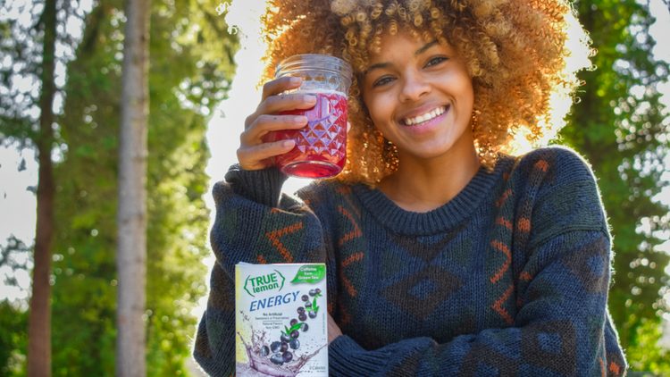 A young woman sits outside in the sun and drinks True Lemon Energy Blueberry Acai, made with Clean Caffeine from green tea. 