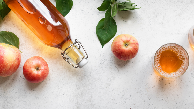 A bottle of Apple Cider Vinegar on a kitchen counter surrounded by apples and glass of the vinegar. 