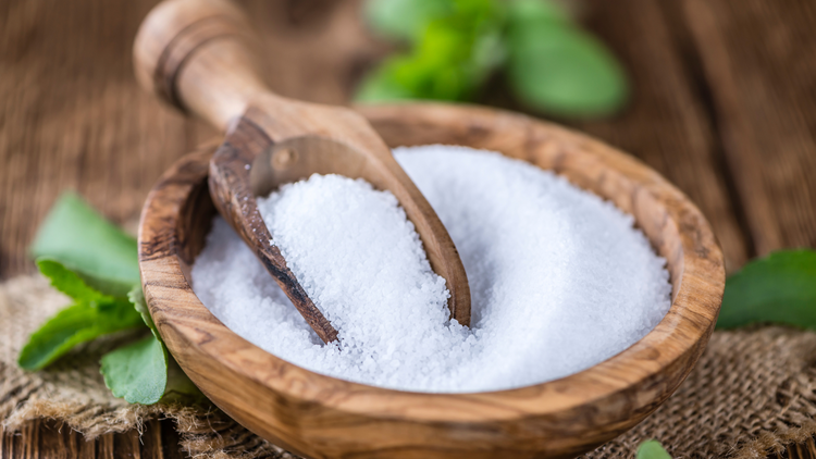 An image of a wooden bowl filled with granules of stevia. A wooden scoop is within the bowl. Behind the bowl are some leaves of the plant that stevia comes from.