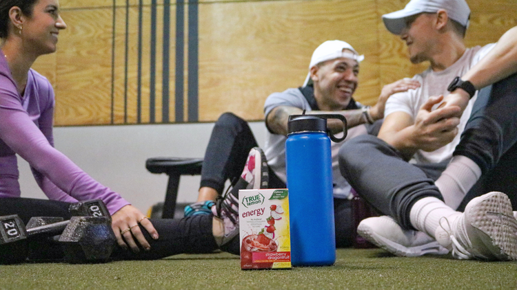 A group of three friends sit on the floor of the gym after a workout and drink True Lemon Energy. 