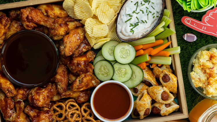 An image of a table with a collection of food. It is setup for a Super Bowl Party, there are wings, dips, chips, cucumbers, carrots, and celery among other things.