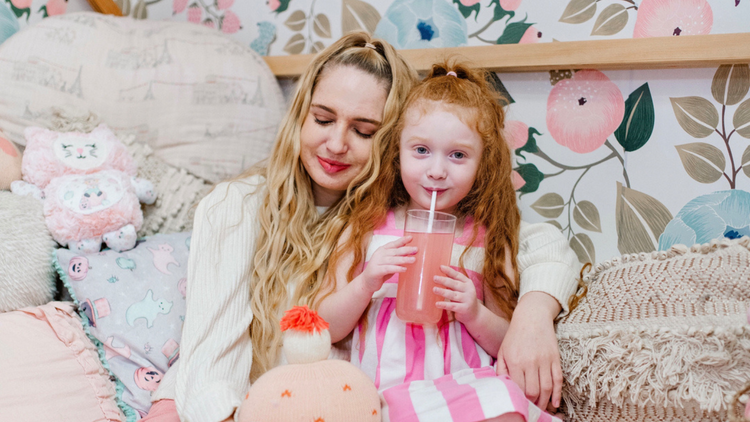 A woman and a little girl are sitting on a bed, the girl is drinking a glass of True Lemon Kids.