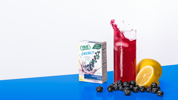 Glass of True Lemon Blueberry Acai drink, lemons, and blueberries, and a box of the Blueberry Acai flavor packets on a bright blue table.
