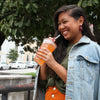 A woman smiles widely and takes a sip of True Orange Mango Orangeade. 