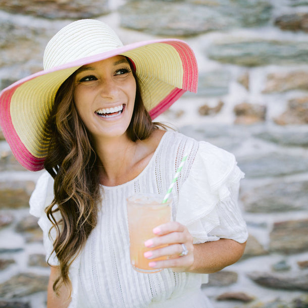 A woman wearing a sunhat and a white blouse laughs and enjoys a glass of True Lemon Peach Lemonade.