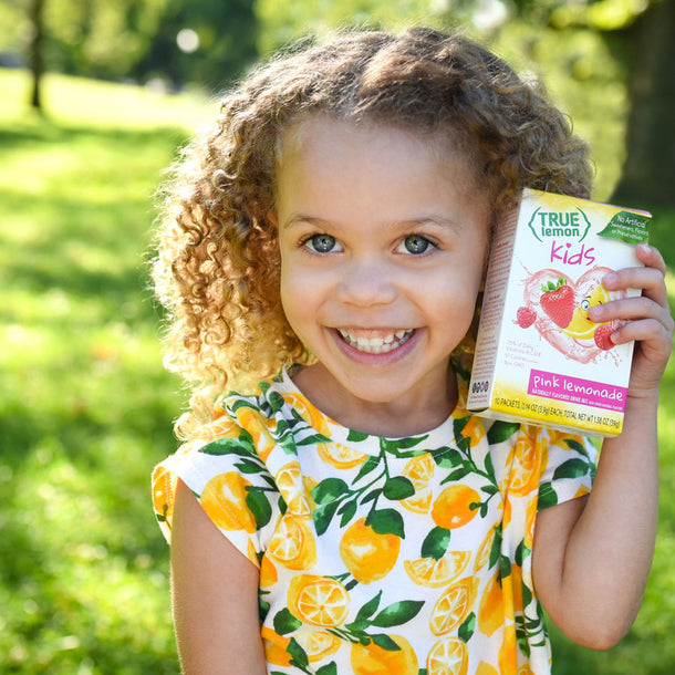 A little girl outside wearing a dress covered in lemons, hold a box of True Lemon Kids Pink Lemonade up to her head.