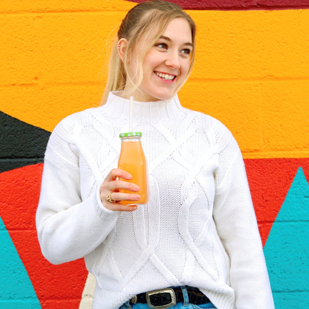 A woman in front of a wall painted with bright orange, red, and blue, smiles and enjoys a bottle of True Orange Mango Orangeade.