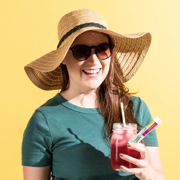 A woman wearing a sunhat and sunglasses smiles widely and drinks a glass of True Lemon Wildberry Lemonade.
