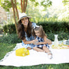 A mom and her daughter picnic outside on a white blanket. The mom gives her daughter a sip of her True Lemon Original Lemonade.